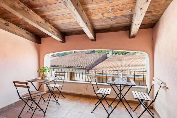 A balcony with wooden beams, a table, and chairs, overlooking a tiled roof.