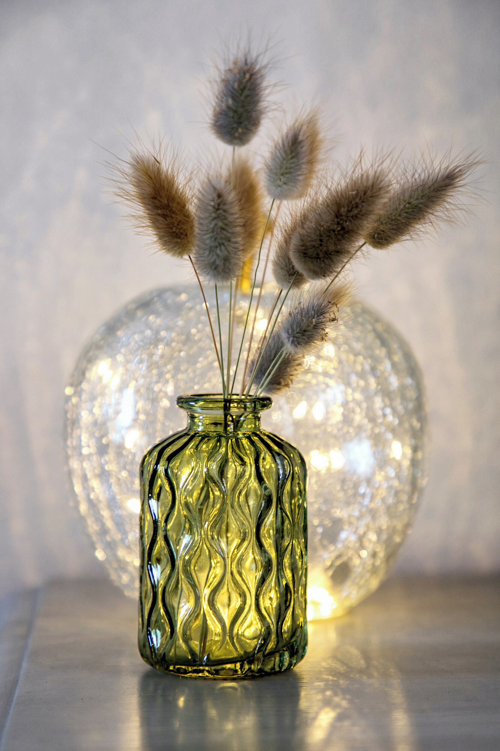 Un vase en verre transparent orné d’un motif ondulé, contenant des herbes de la pampa séchées, placé sur un fond légèrement éclairé avec un objet sphérique.