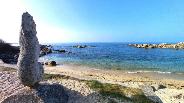 Paisaje costero con una formación rocosa destacada en primer plano, una playa arenosa y aguas marinas tranquilas.