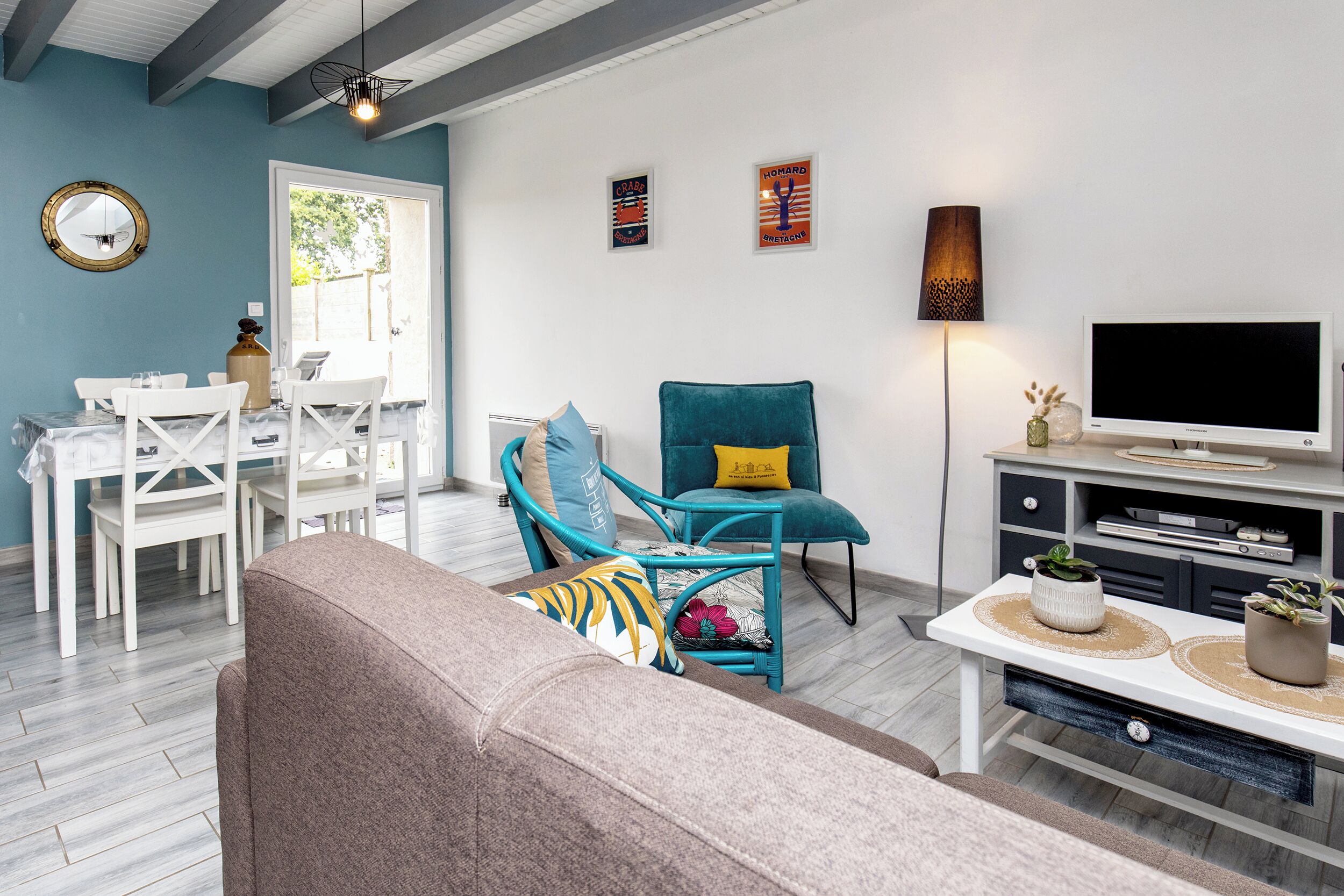 Un salon comprenant un canapé, un fauteuil turquoise, un meuble pour la télévision et un coin repas avec une table et des chaises blanches.