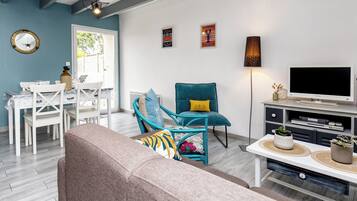 Un salon comprenant un canapé, un fauteuil turquoise, un meuble pour la télévision et un coin repas avec une table et des chaises blanches.