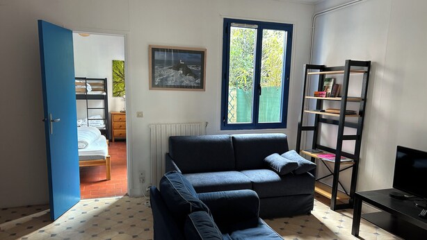 Ein Wohnzimmer mit blauem Sofa, Fernseher, Bücherregal und einem Fenster mit Blick auf Grünfläche.