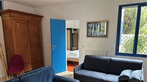 Ein Zimmer mit blauer T ür, einem Holzschrank, einem grauen Sofa, einem roten Sessel und einem Fenster mit Blick ins Grüne.