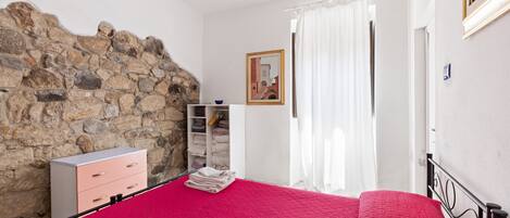 Un dormitorio con pared de piedra, una cama con colcha roja, un aparador, un armario y un cuadro en la pared.