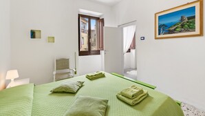A bedroom with a bed, pillows, a painting, a window, and a door leading to another room.