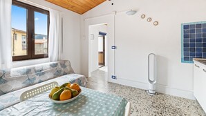 A room with a wooden ceiling, a bed with a polka dot bedspread, a bowl of fruit on the bed, a white radiator, and a view of the outside through a window.