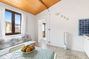 A room with a wooden ceiling, a bed with a polka dot bedspread, a bowl of fruit on the bed, a white radiator, and a view of the outside through a window.