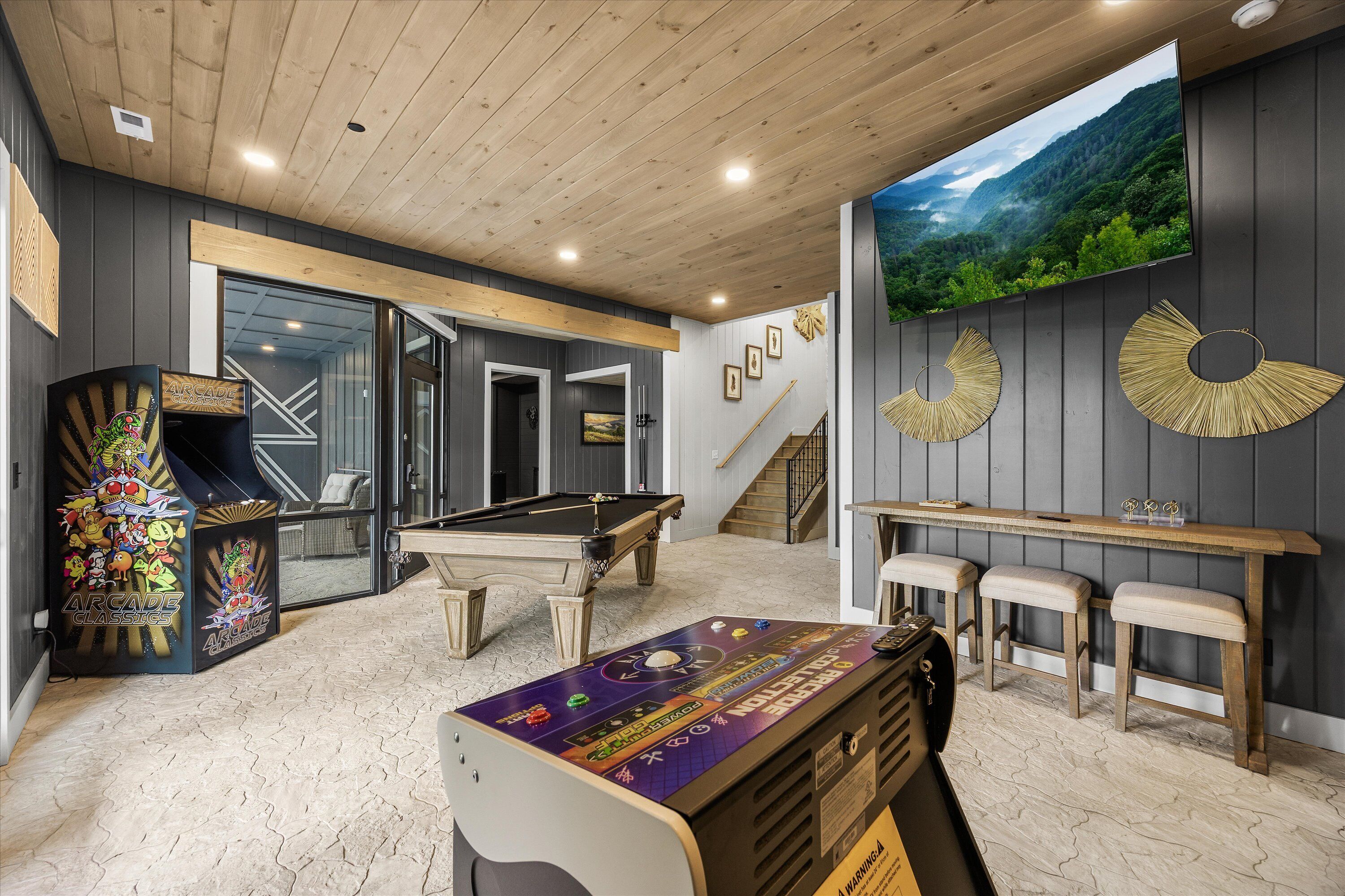 A pool table in a room with a wooden ceiling, a large TV screen, and a bar area with stools.