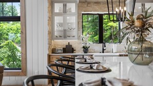 A modern kitchen with a large window, white cabinetry, and a dining area with chairs and a table set for two.