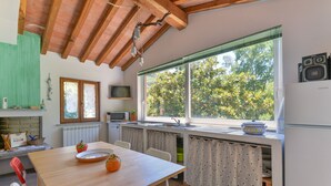 A kitchen with a wooden table, white chairs, a window with a view of trees, a sink, and a refrigerator. - Villa 'I Fiori Redinoce' with Private Terrace, Private Garden, and Air Conditioning (Procchio)
