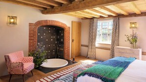A bedroom with a brick fireplace, a wooden ceiling, a bed with a colorful blanket, a chair, and a window with patterned curtains. - Wilderness Reserve - The Grange (Halesworth)