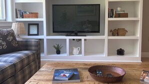 Een woonkamer met een flatscreen televisie, een bank, een bijzettafeltje met boeken en een schaal, en witte planken met decoratieve objecten.