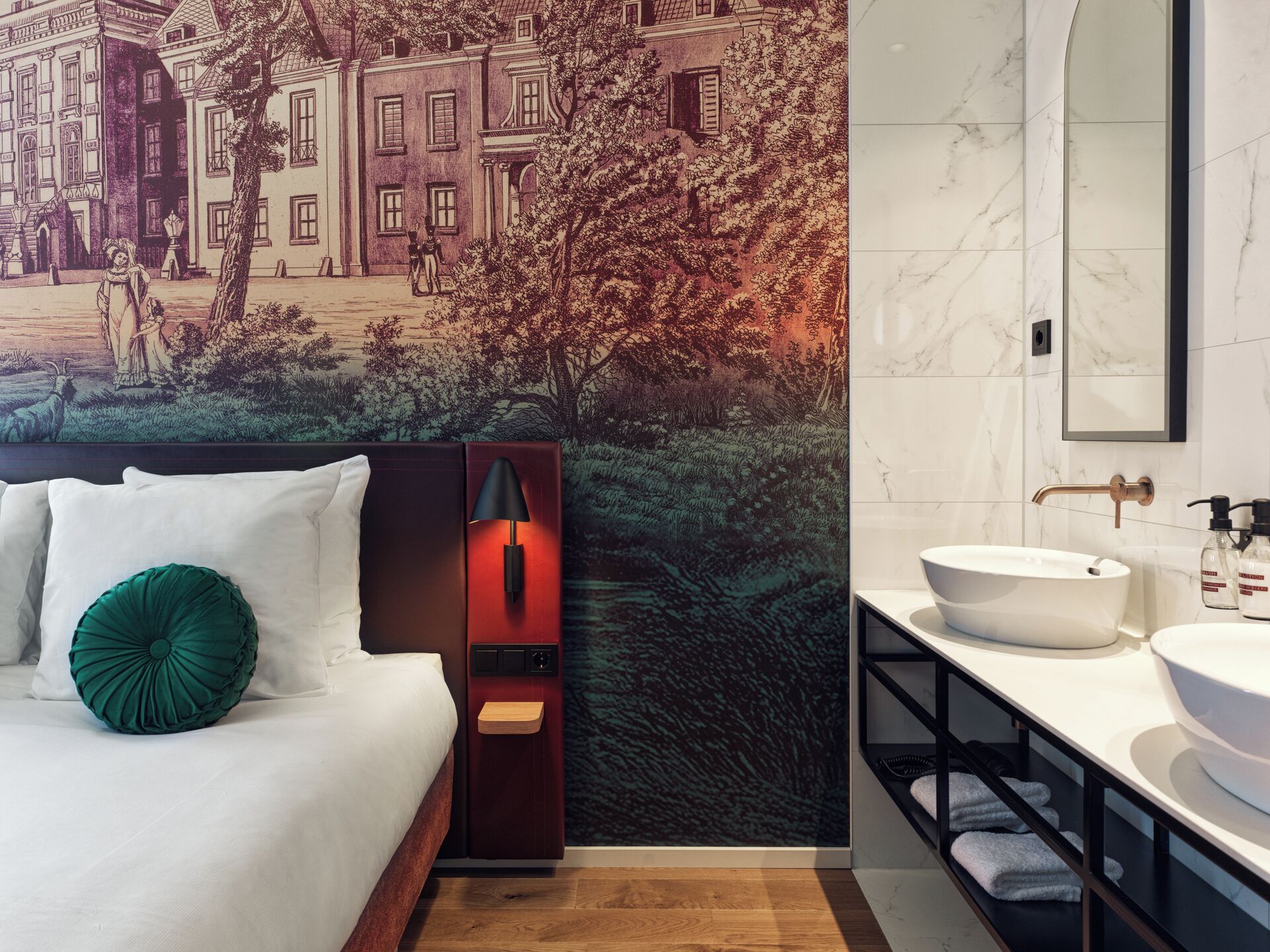 Quarto de hotel moderno com uma cama grande, uma mesinha de cabeceira com abajur, e um banheiro com um espelho amplo e uma pia.