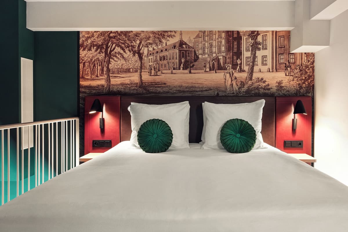 A bedroom with a large bed, two green decorative pillows, a headboard with wall-mounted lights, and a cityscape mural above the bed.