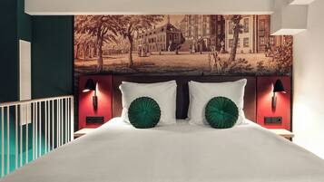 Une chambre à coucher avec un grand lit, deux coussins décoratifs verts, une tête de lit dotée de lampes murales et une fresque représentant un paysage urbain au-dessus du lit.