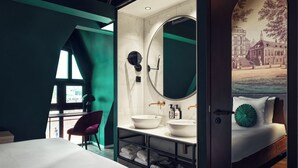 Ein modernes Hotelzimmer mit einem großen Bett, einem Schminktisch mit rundem Spiegel und Blick auf eine Stadtlandschaft durch ein Fenster.