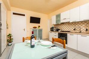 A modern kitchen with white cabinets, a flat-screen TV, a dining table with chairs, and a wine bottle.