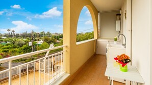 Un balcón con vistas a zonas verdes y edificios, una pequeña cocina con un lavabo y un jarrón de flores.