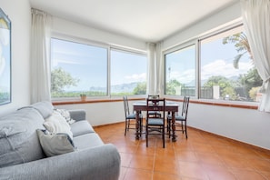A room with a sofa, dining table, chairs, large windows, and a view of greenery and mountains.