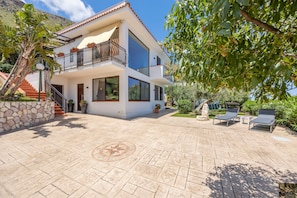 A two-story house with a balcony, a stone wall, and a paved patio with lounge chairs.