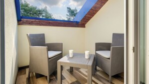 Un petit balcon avec des chaises et une table en osier, deux tasses blanches, et une vue sur les arbres à travers le puits de lumière.