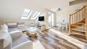 Un salon moderne avec un canapé, une table basse et un coin repas comprenant une table et des chaises. Un téléviseur est fixé au mur, il y a un ventilateur de plafond et un escalier.