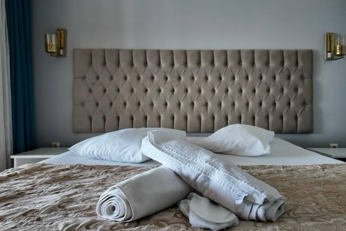 A neatly made bed with a tufted headboard, white pillows, and rolled towels on a patterned bedspread.