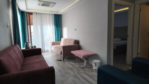 Ένα σύγχρονο δωμάτιο ξενοδοχείου με έναν κόκκινο καναπέ, μια ροζ πολυθρόνα, ένα μικρό τραπέζι με ένα τραπεζομάντιλο με ριγέ σχέδιο και ένα κρεβάτι στο βάθος.