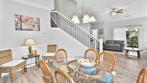 A dining area with a glass table surrounded by wicker chairs, a staircase, and a gray sofa in the background. - Charming Townhome in Barefoot Resort — Minutes from Beach, Golf & Fun! (N. Myrtle Beach)
