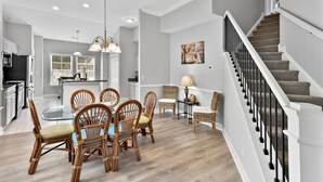 A modern kitchen with white cabinets, a dining area with wicker chairs, and a staircase with a black railing. - Charming Townhome in Barefoot Resort — Minutes from Beach, Golf & Fun! (N. Myrtle Beach)