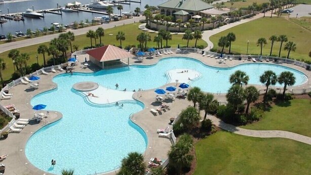 A large, kidney-shaped swimming pool with multiple lanes, surrounded by lounge chairs and umbrellas. - Charming Townhome in Barefoot Resort — Minutes from Beach, Golf & Fun! (N. Myrtle Beach)
