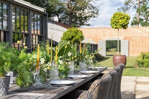 An outdoor dining area with a long wooden table set for a meal, surrounded by potted plants and candles.