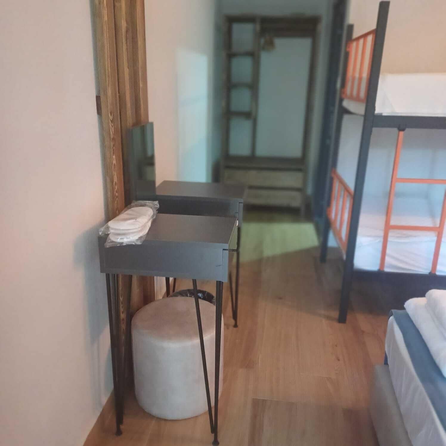 A room with a bunk bed, a desk with a chair, a small stool, and a mirror on the wall.