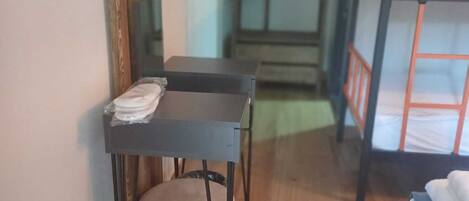 A room with a bunk bed, a desk with a chair, a small stool, and a mirror on the wall.