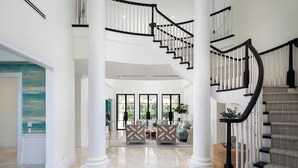 A spacious foyer with a grand staircase, a central seating area, and large windows allowing natural light. - Port Royal - Sweeping Lake Views (Naples)