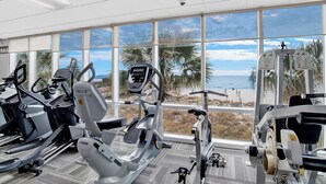 Ein Fitnessstudio mit verschiedenen Trainingsgeräten, großen Fenstern und Blick auf den Strand und Palmen.