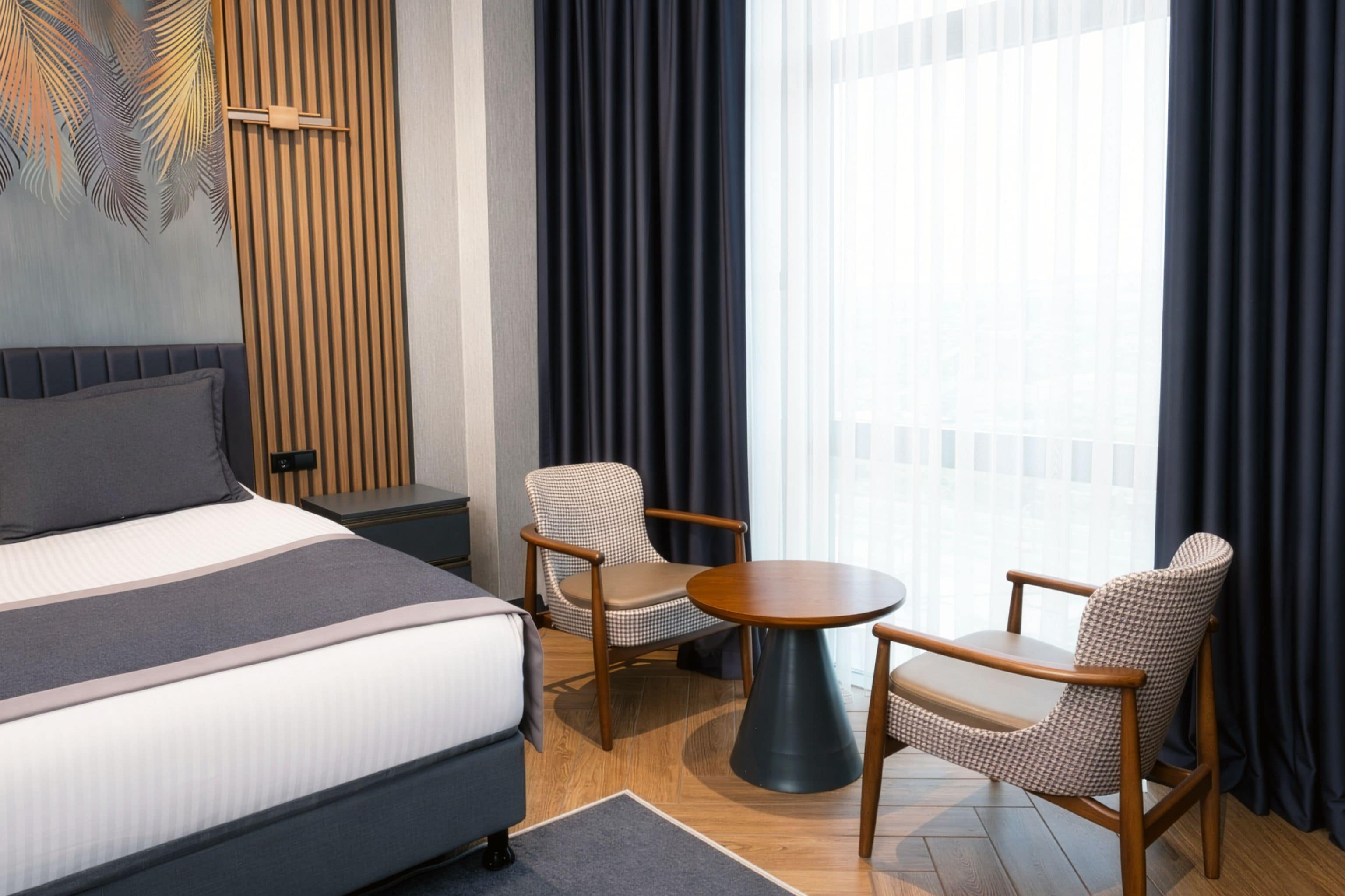 A hotel room with a bed, two armchairs, a small round table, and a view of the city through large windows.