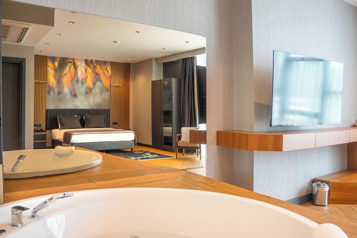 A modern hotel room with a large bed, a sitting area, a large window, and a television mounted on the wall.