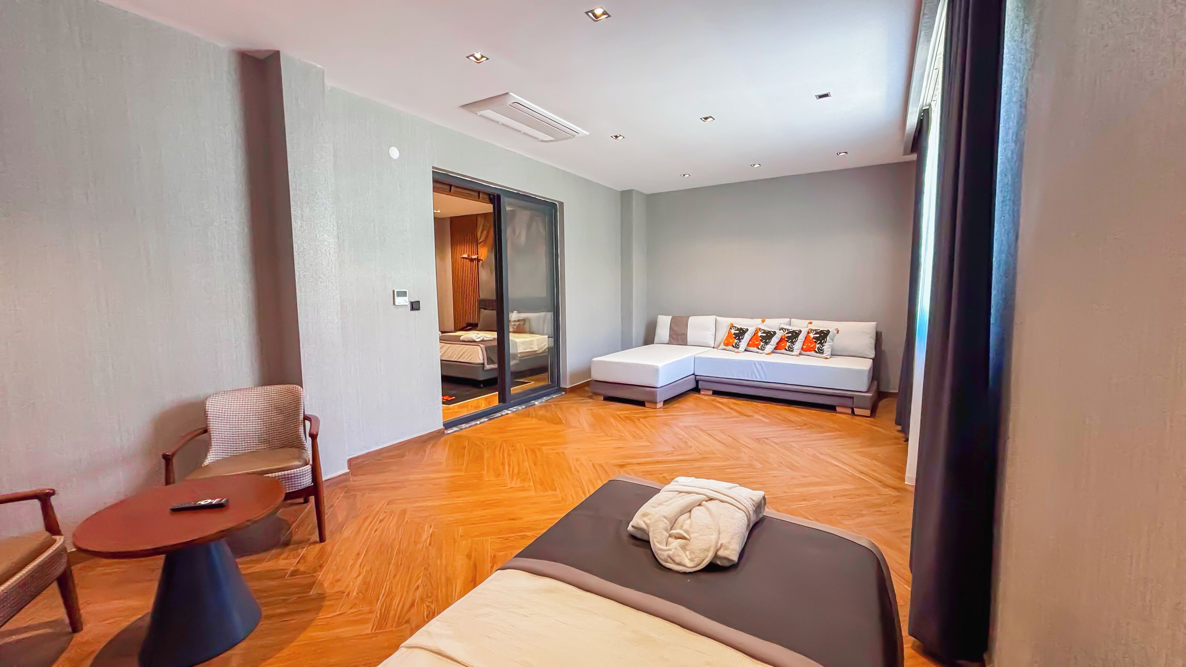 A modern hotel room with a wooden floor, a sofa bed with pillows, a small table with a remote control, and a chair.