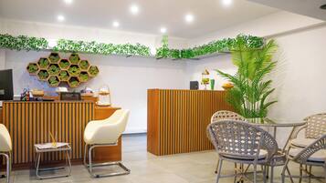 A reception area with a wooden reception desk, two white chairs, wicker chairs around a small table, and a large potted plant.