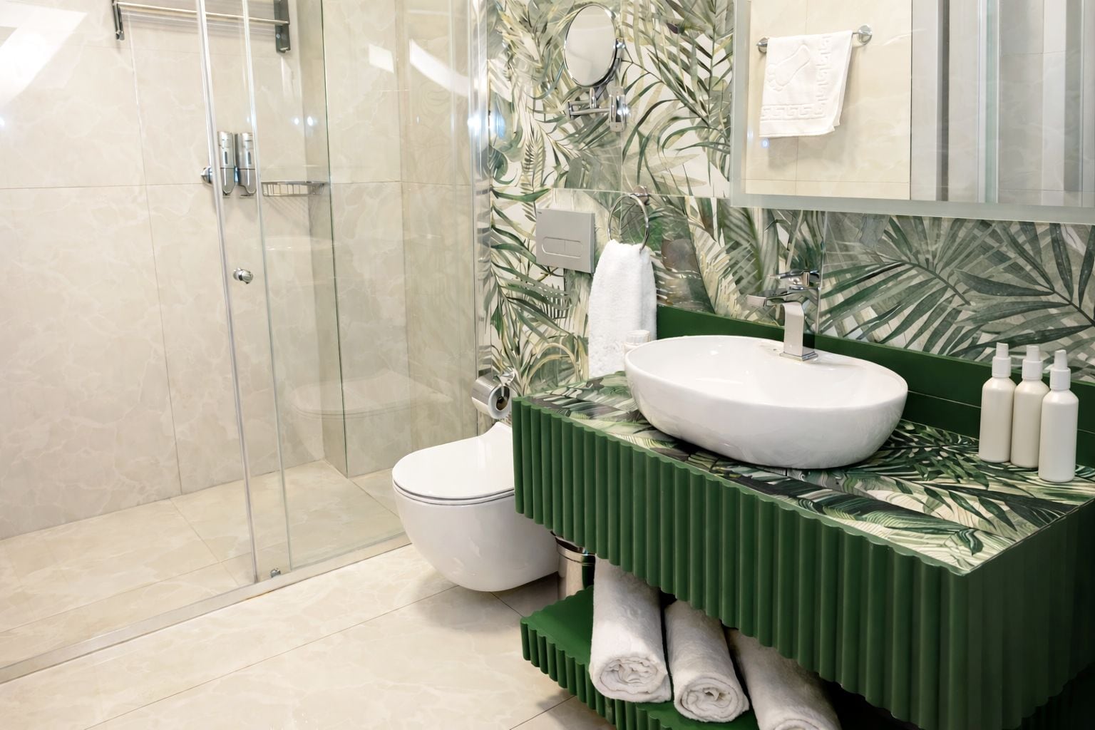 Un baño moderno con cabina de ducha de vidrio, inodoro blanco, tocador verde con lavabo redondo y espejo con fondo de motivos vegetales.