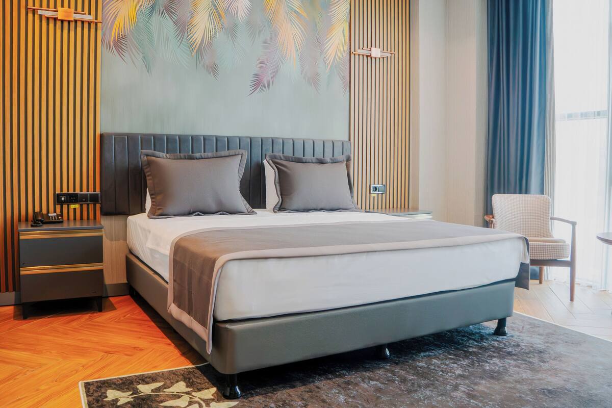 A modern bedroom with a large bed, a nightstand, a chair, and a wall with a palm leaf mural.