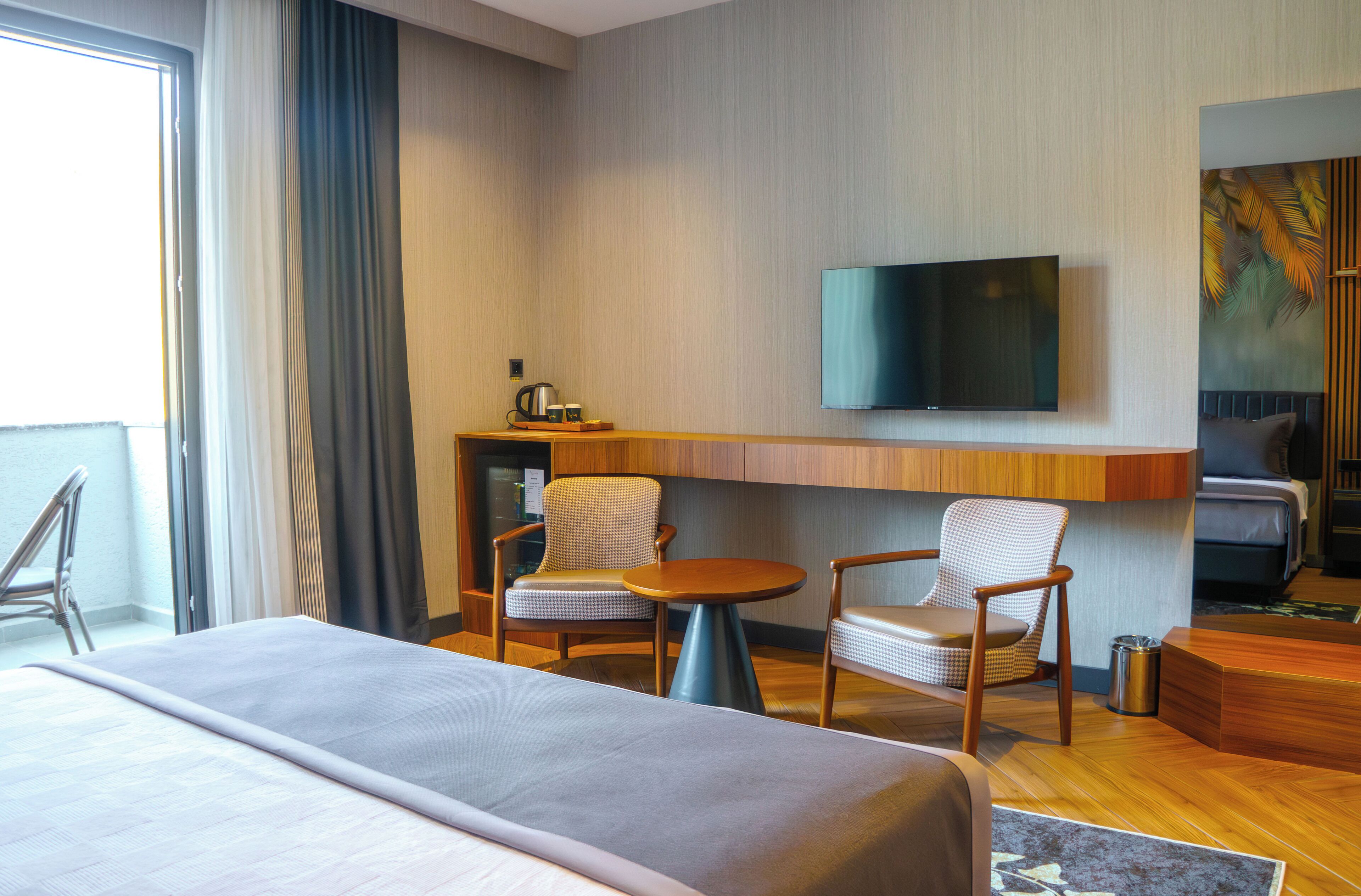 A hotel room with a bed, two chairs, a small table, a TV mounted on the wall, and a large window with curtains.