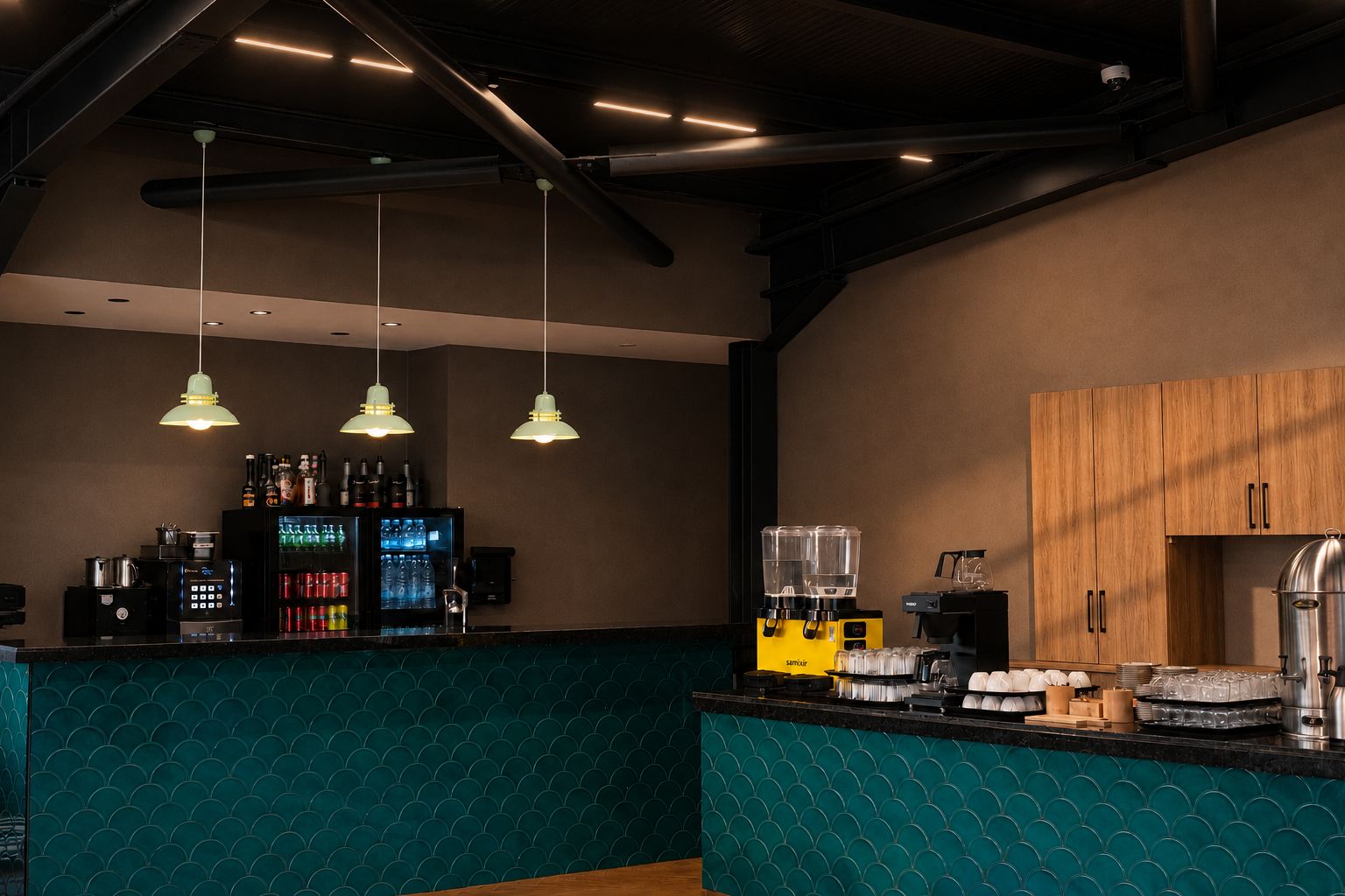 Una zona de bar con pared de color turquesa con diseño, lámparas colgantes, un refrigerador lleno de bebidas y una máquina de café.