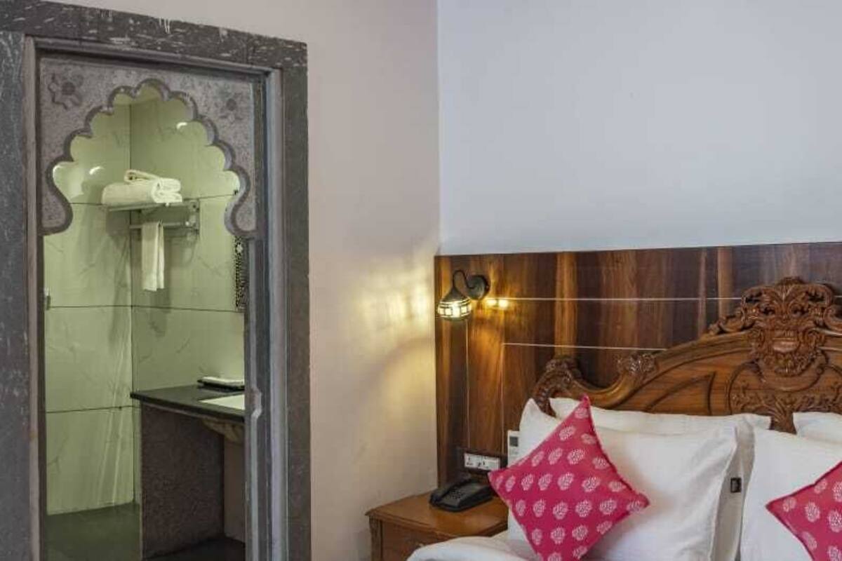 A hotel room with a wooden headboard, two white pillows with pink patterns, a nightstand with a phone, and a mirror with a decorative frame.