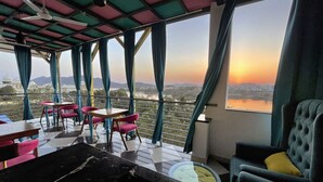 A dining area with a table and chairs, large windows with curtains, and a view of a landscape with mountains and a body of water. - Aaj Haveli - A Luxury Boutique Hotel (Udaipur)