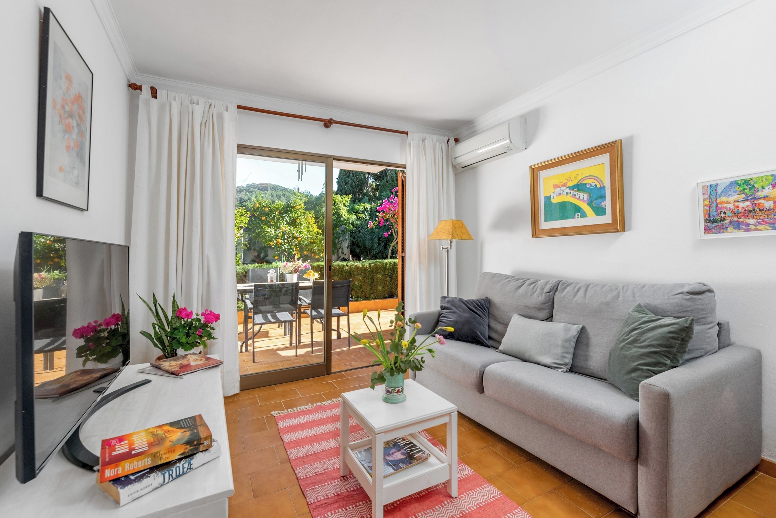 Uma sala de estar moderna com um sofá cinzento, uma TV de tela plana, uma pequena mesa branca com um vaso de flores e um tapete vermelho.