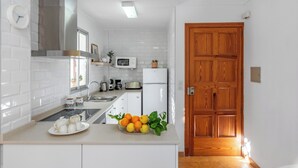 A modern kitchen with white cabinetry, a stainless steel range hood, a microwave, and a refrigerator. There's a wooden door to the right and a small window above the sink. - Apartment Bougainvillea,  a beautiful apartment just a few steps from the beach. (Port de Sóller)