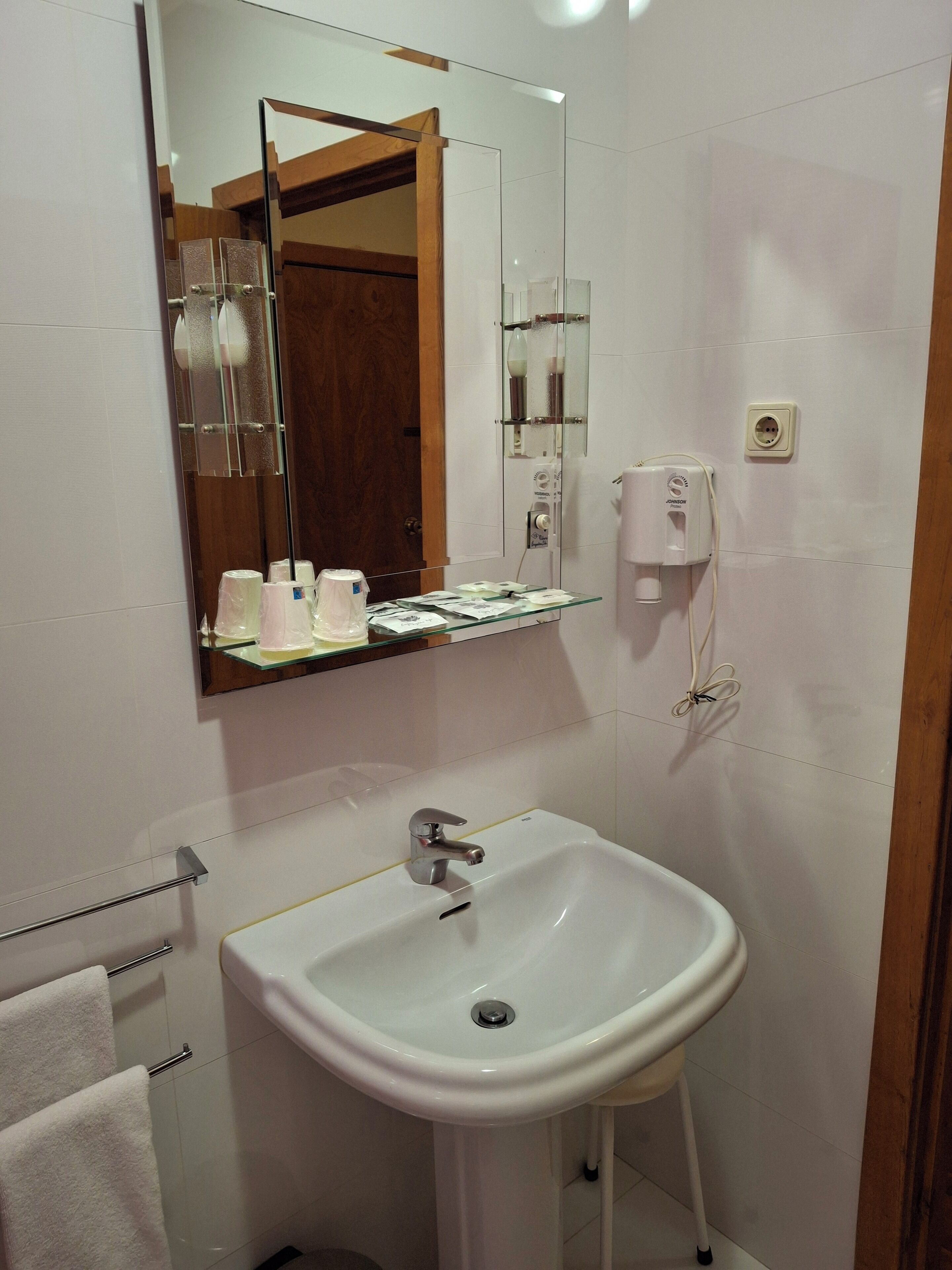 Une salle de bain avec un lavabo blanc, un miroir, un porte-serviettes et une étagère en verre où sont rangés les articles de toilette.