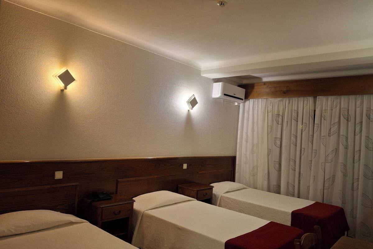 A hotel room with two single beds, a wooden headboard, a nightstand with a phone, a wall-mounted air conditioner, and a patterned curtain.